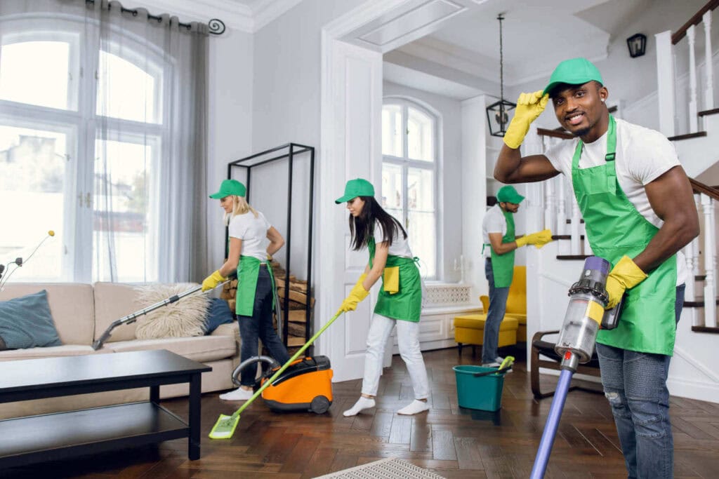 Four people in green aprons, caps, and yellow gloves are cleaning a modern, bright living room with various tools. This NY Cleaning Services Long Island team works efficiently in a neat space with large windows and stylish furniture.