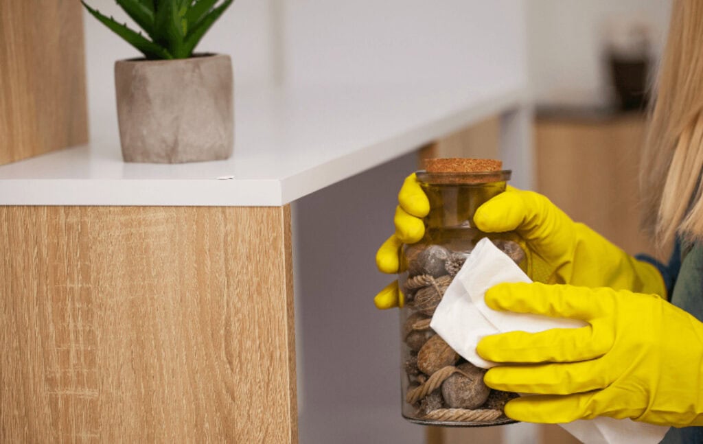A person wearing yellow rubber gloves cleans a glass jar filled with shells using a white cloth. In the background, a potted plant sits on a wooden shelf—a scene reminiscent of Cleaning Services Long Island, NY standards.