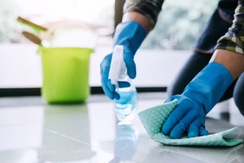 A person wearing blue gloves cleans a shiny floor with a green cloth and spray bottle—typical of NY Cleaning Services Long Island. A green bucket with cleaning supplies is visible in the blurred background.