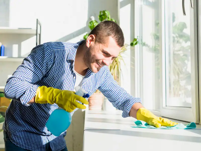 A man wearing yellow gloves and a blue checkered shirt cleans a windowsill with a spray bottle and cloth, smiling in a bright, sunlit room—just another day for top Cleaning Services Long Island, NY, with plants in the background.