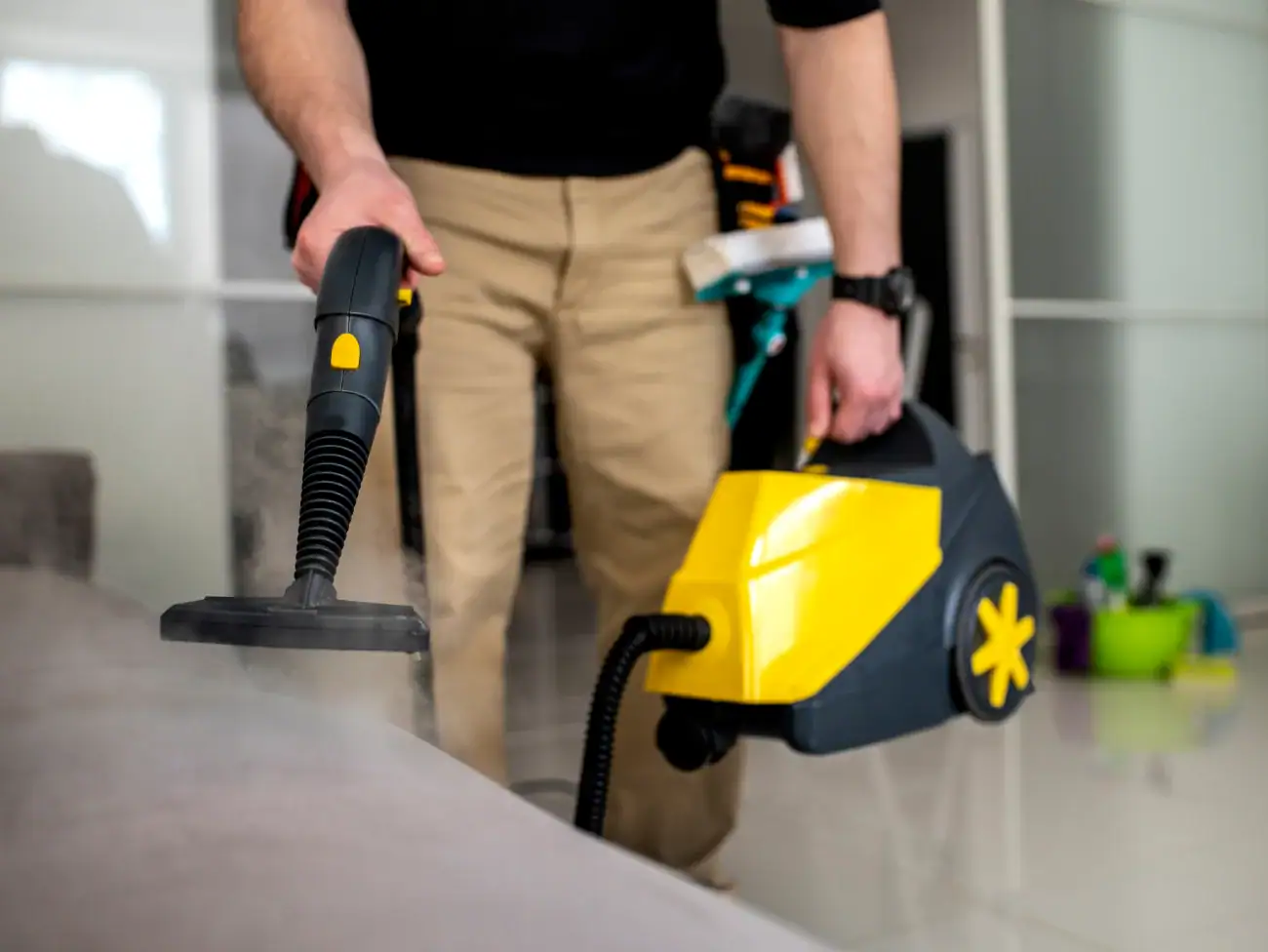 A person using a yellow and black steam cleaner to clean a sofa in a modern, bright living room, showcasing professional Cleaning Services Long Island, NY. The focus is on the device and the person’s hands.