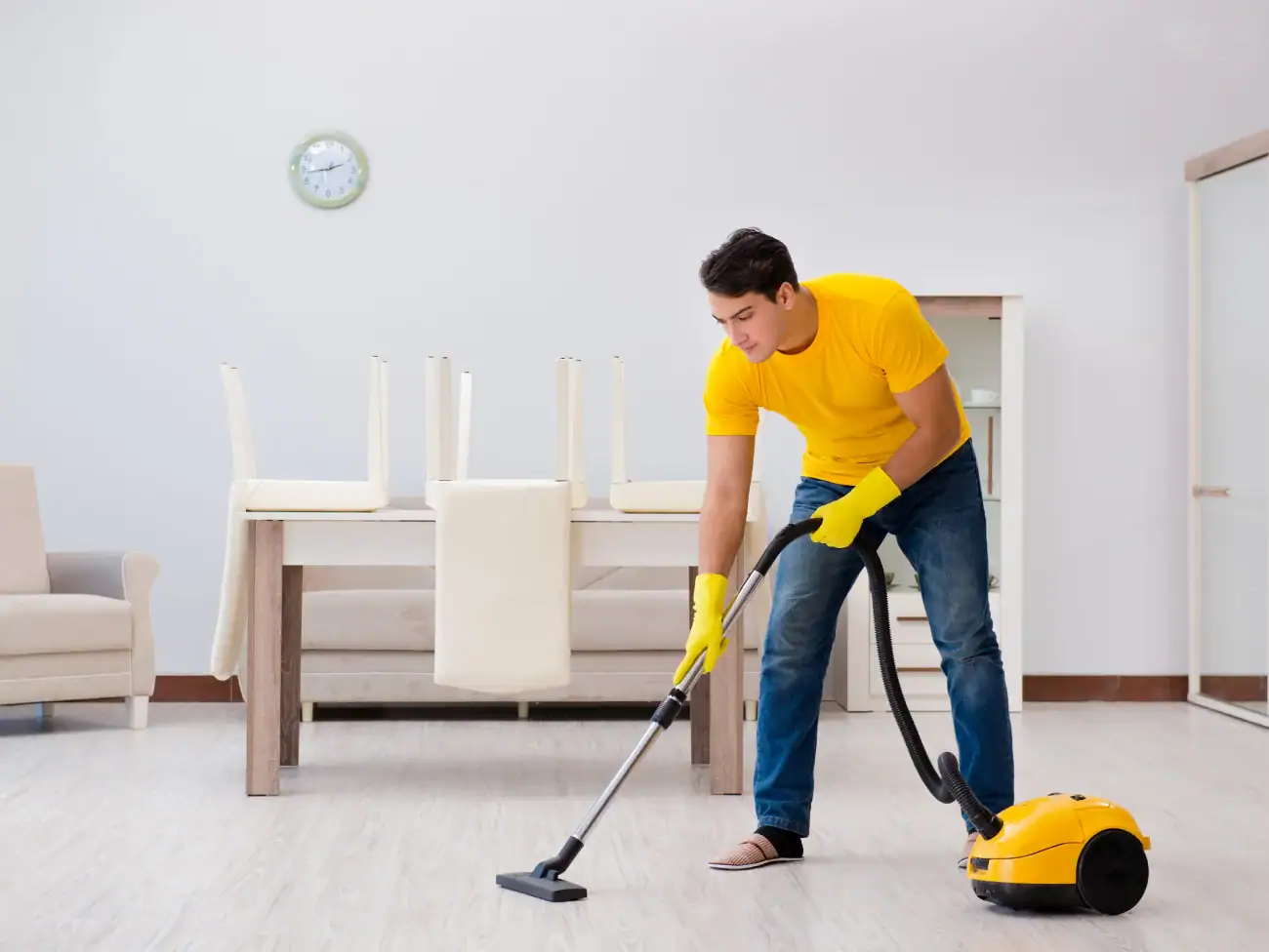 A man wearing a yellow shirt and gloves vacuums the floor in a bright, tidy room—showcasing the high standards of NY Cleaning Services Long Island. Dining chairs are stacked on the table, and a clock hangs on the white wall above.
