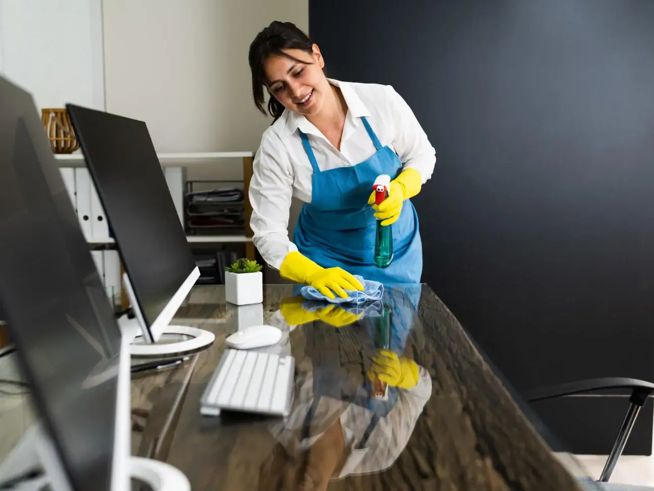 A woman wearing an apron and yellow gloves smiles while cleaning a desk in an office, representing professional Cleaning Services Long Island, NY. She uses a spray bottle and cloth near computer monitors, a keyboard, mouse, and small plant.