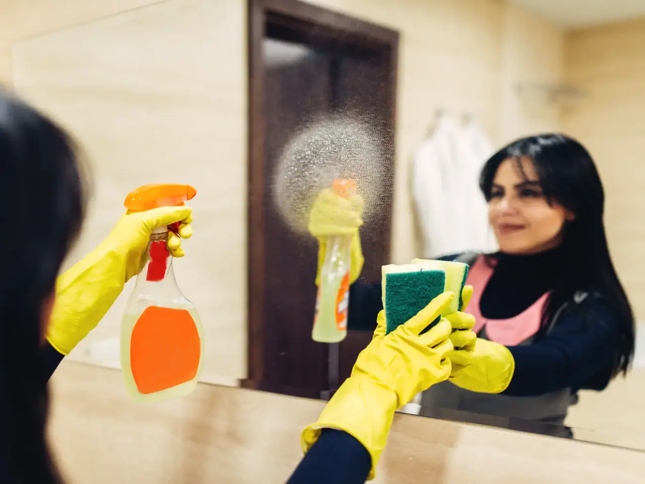 A person wearing yellow gloves cleans a mirror with a spray bottle and sponge in a bathroom, their reflection visible—just like the attention to detail offered by Cleaning Services Long Island, NY.