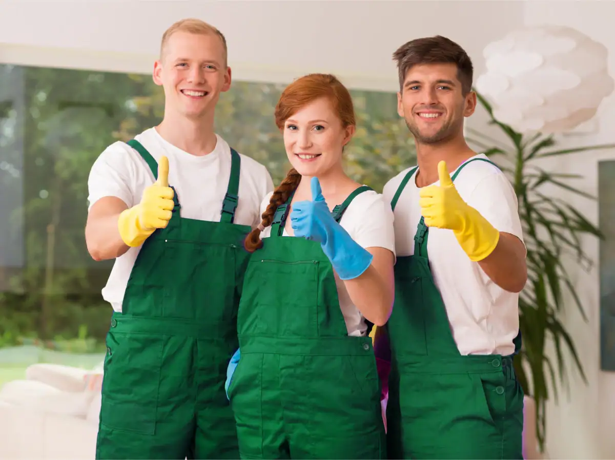 Three smiling people in green overalls and rubber gloves stand indoors, giving thumbs up to the camera, suggesting they're part of a Cleaning Services Long Island crew ready for work in NY.