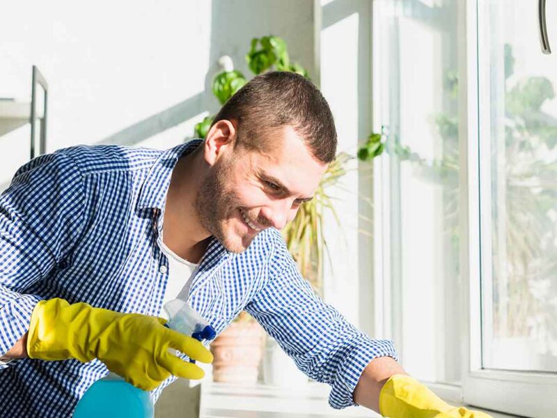 A man wearing yellow rubber gloves and a checkered shirt smiles while cleaning a window with a spray bottle in a bright room, representing top-notch Cleaning Services Long Island, NY, with green plants in the background.