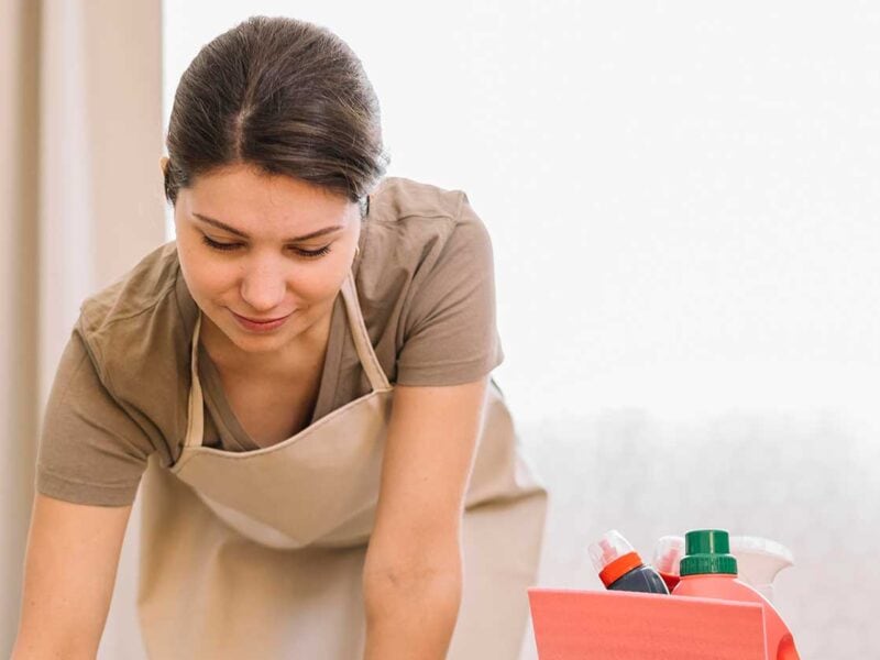 A woman wearing a beige apron is leaning forward while cleaning. A red container with cleaning supplies is beside her. She appears focused on her task, representing Cleaning Services Long Island, NY, with a light background behind her.