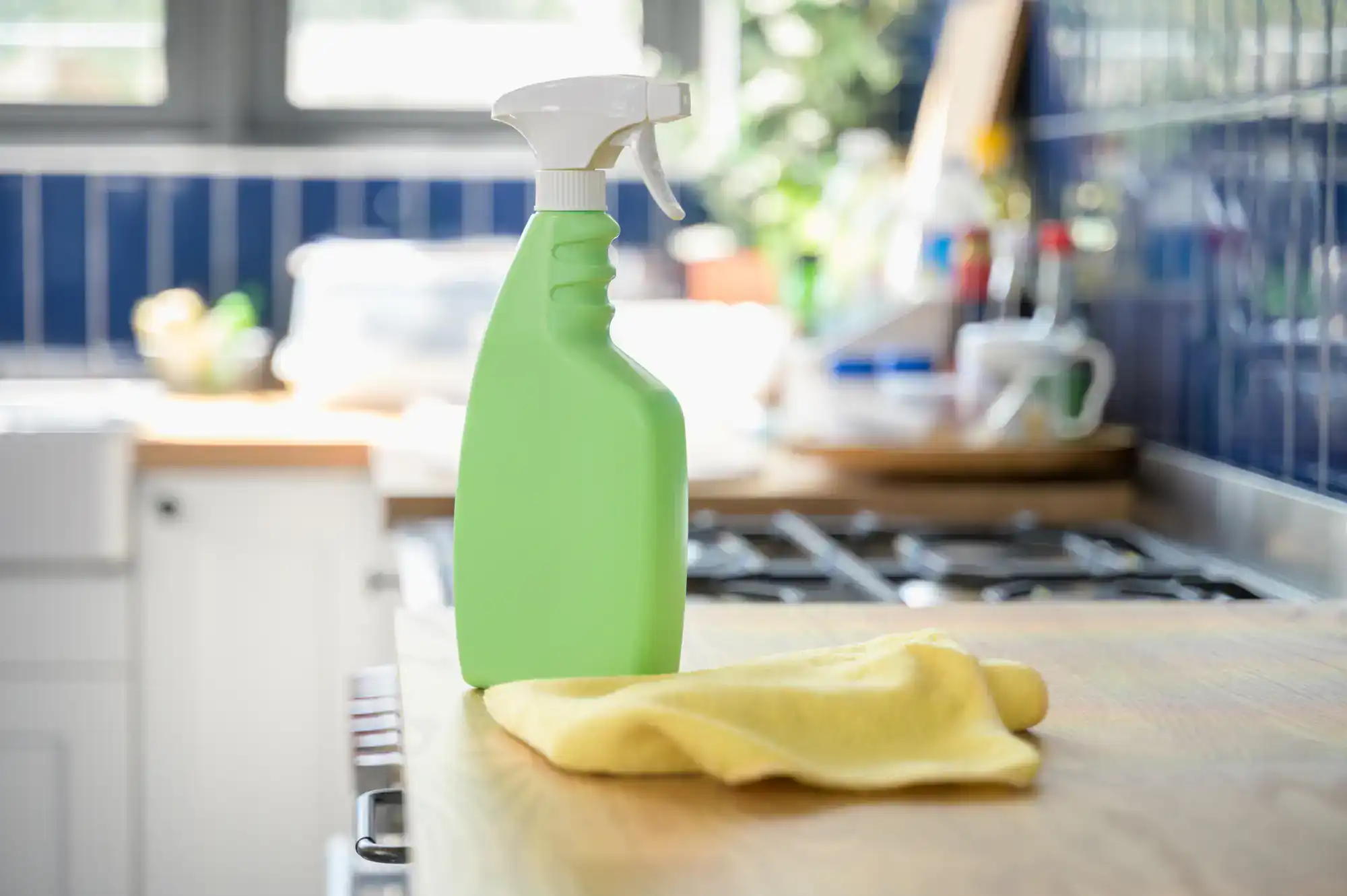 A green spray bottle and a yellow cleaning cloth sit on a wooden kitchen counter, suggesting the fresh impact of Cleaning Services Long Island, NY, with a stove and kitchen items blurred in the background.