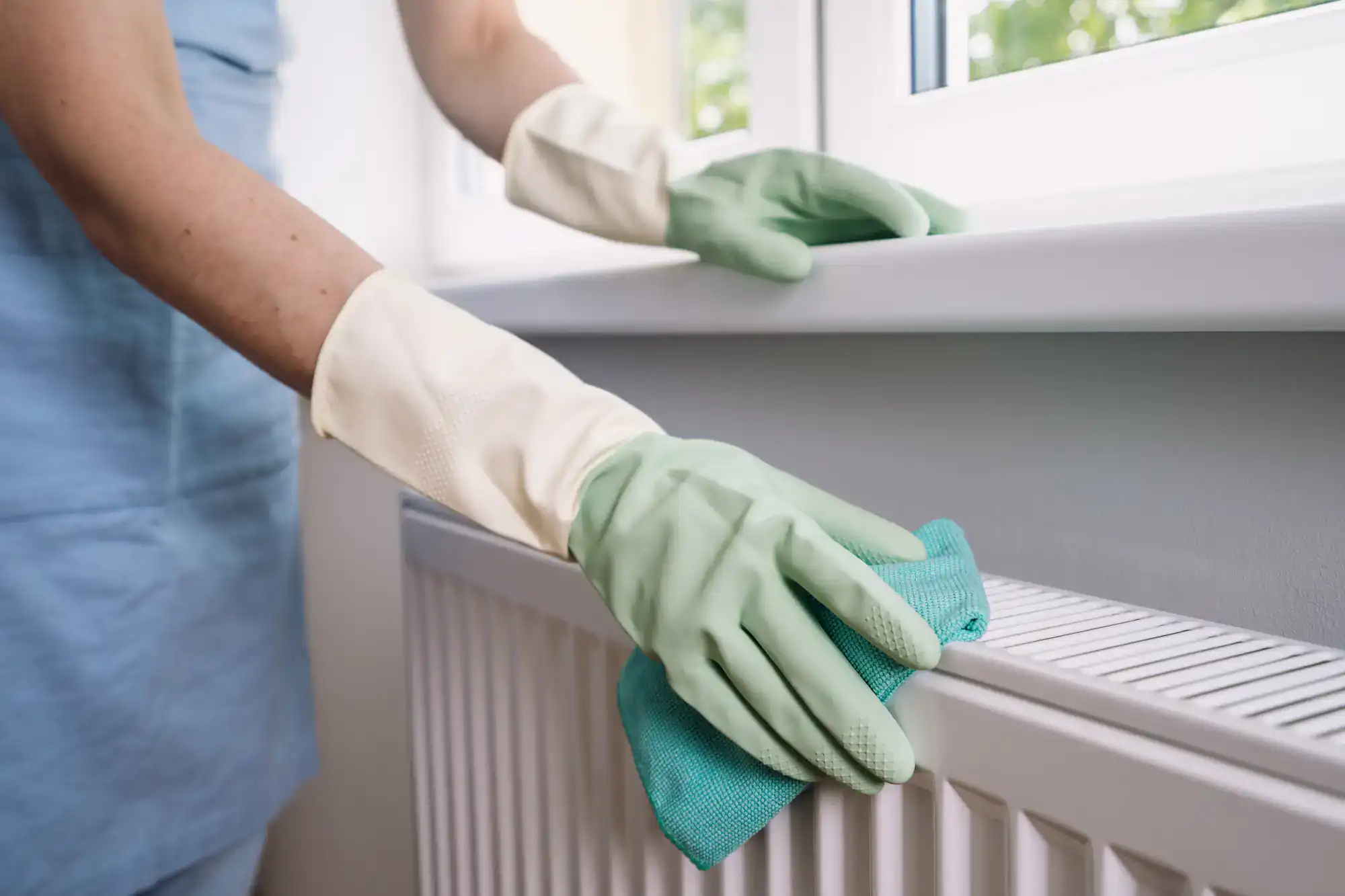 A person wearing green rubber gloves is cleaning a white radiator and window sill with a turquoise cloth. Sunlight streams through the window, reflecting the quality offered by Cleaning Services Long Island, NY.