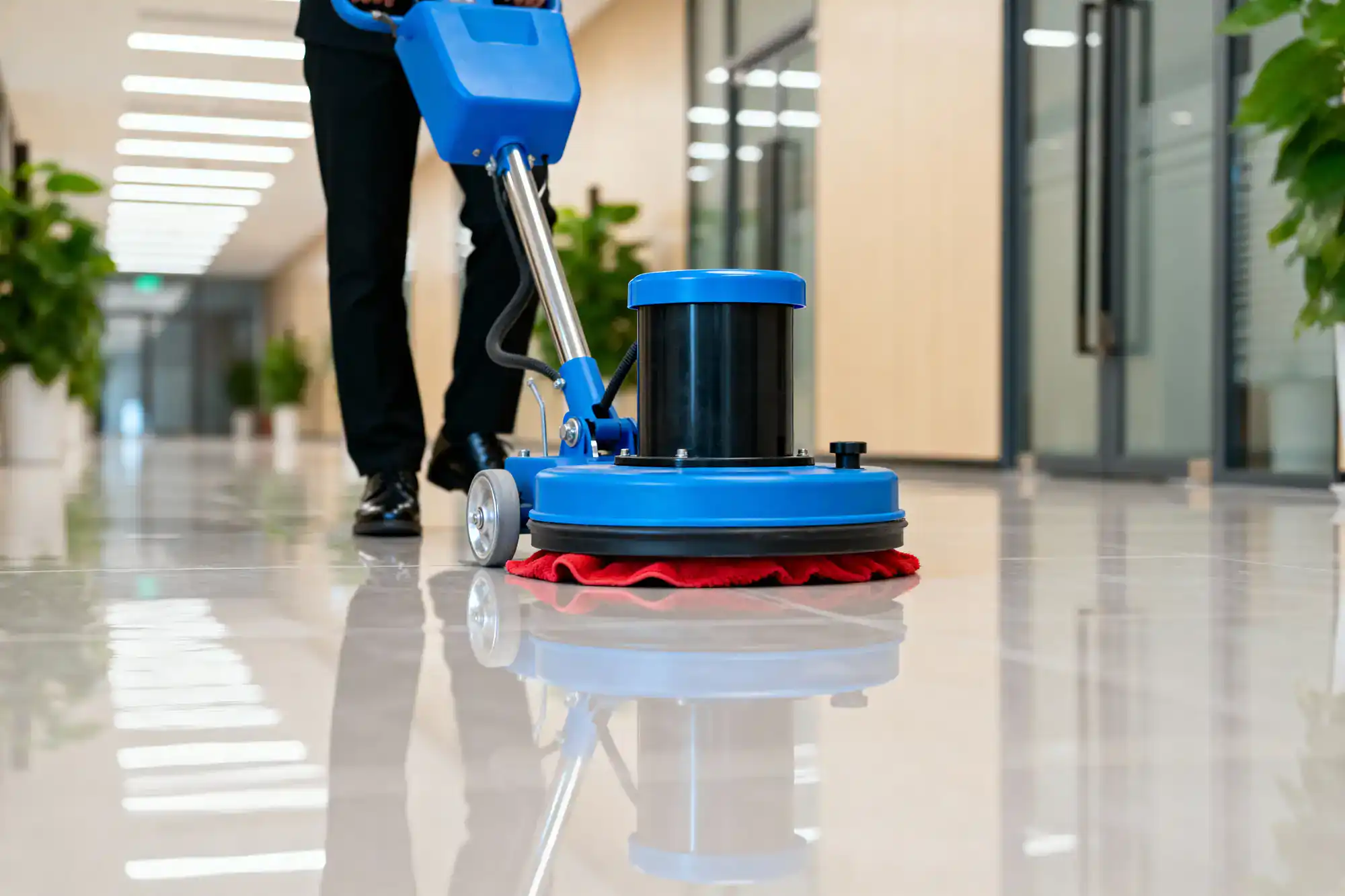 A person operates a blue floor polishing machine on a shiny hallway floor, with green plants and glass office doors in the background, showcasing the quality of Cleaning Services Long Island in NY.