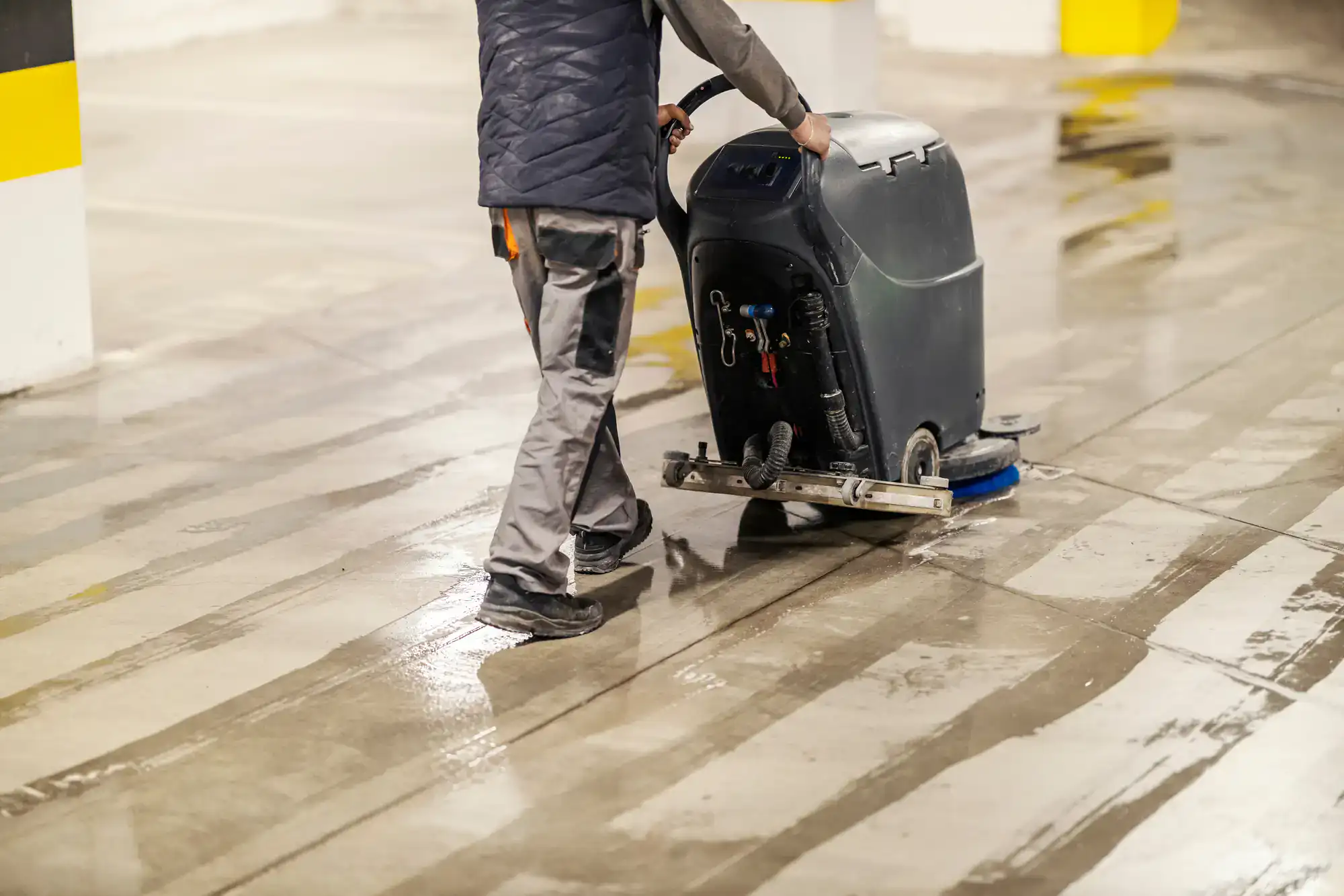 A person from Cleaning Services Long Island operates a large floor cleaning machine on a wet concrete surface in an indoor NY parking garage, wearing work pants and a jacket.