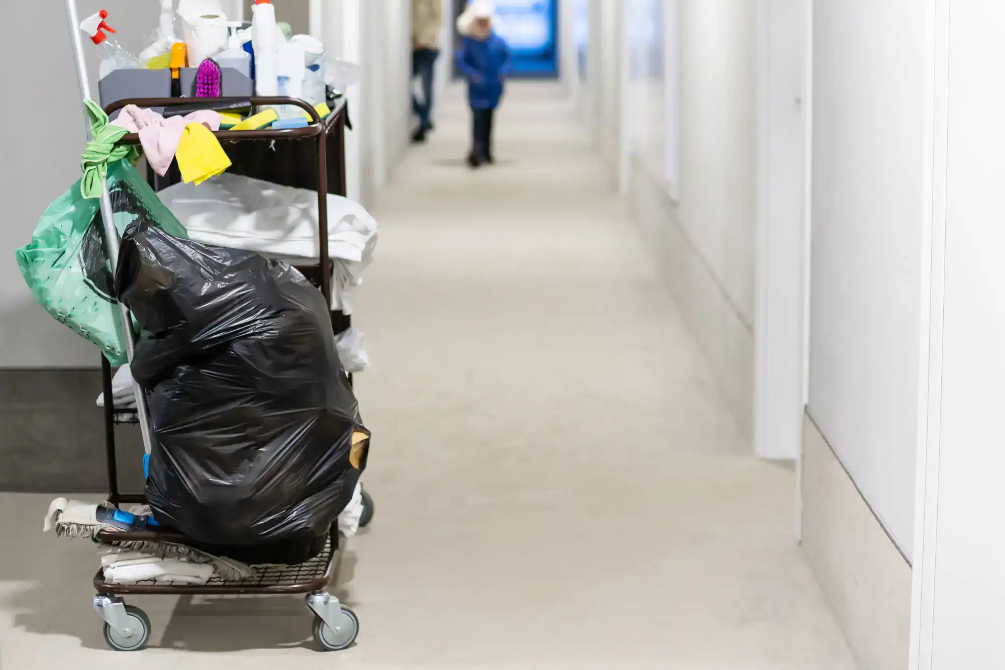 A janitor&rsquo;s cleaning cart with a large black trash bag, cleaning supplies, and rags sits in a hallway; a person and child are visible in the distance&mdash;a typical scene for Cleaning Services Long Island, NY.