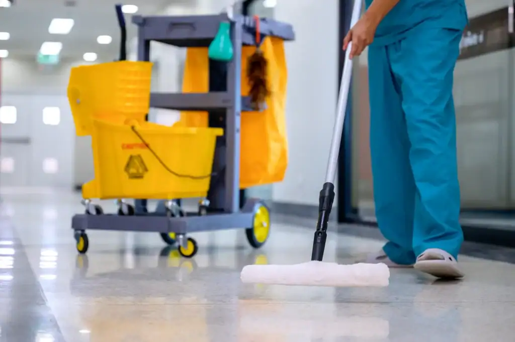 A person in blue scrubs mops a shiny hallway floor. A yellow mop bucket and cleaning cart are visible in the background, showcasing professional Cleaning Services Long Island trusts for spotless NY commercial or hospital settings.