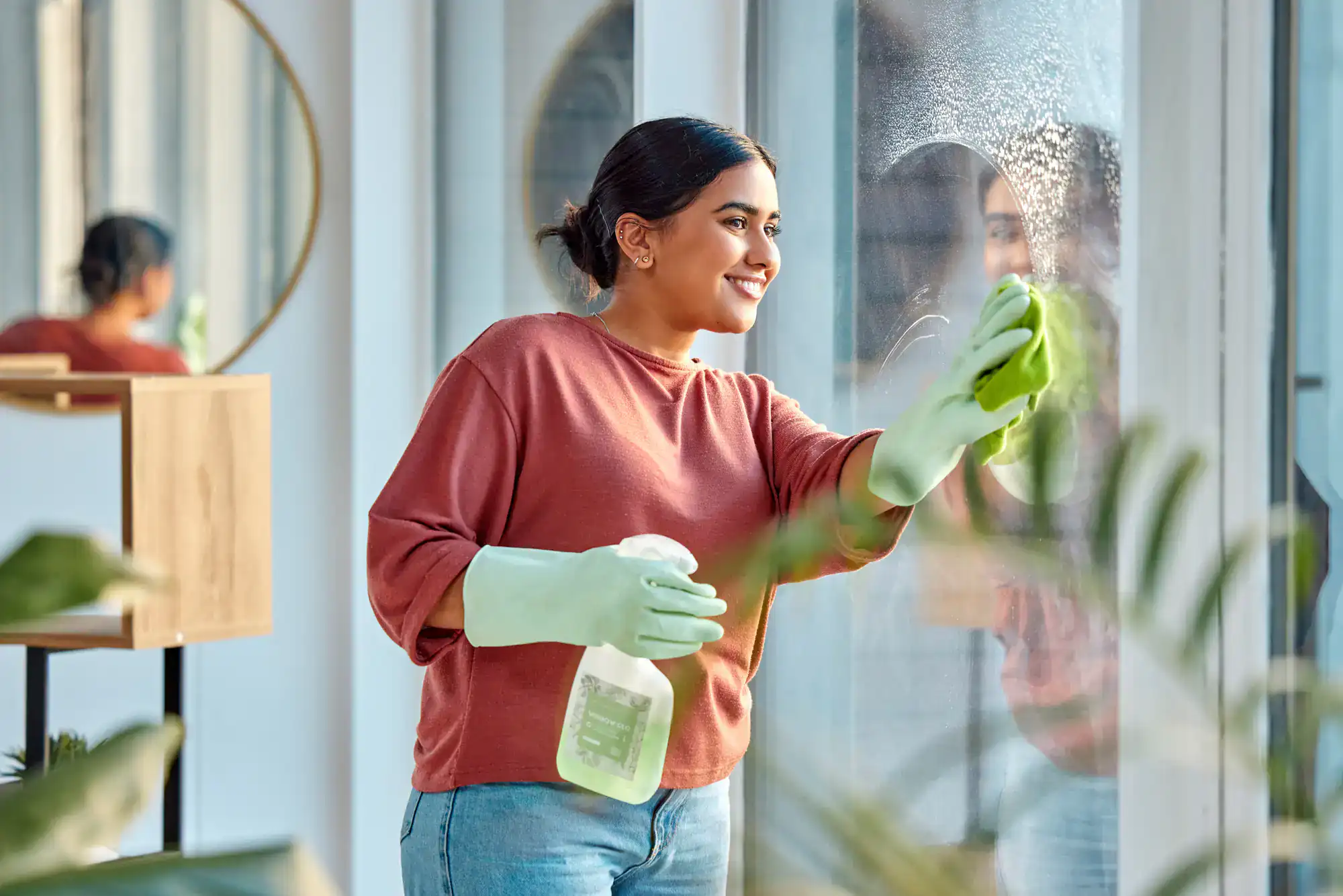 A woman wearing green rubber gloves and a red sweater smiles while cleaning a window with a spray bottle and cloth in a bright room, showcasing professional Cleaning Services Long Island, NY, with plants and a round mirror in the background.