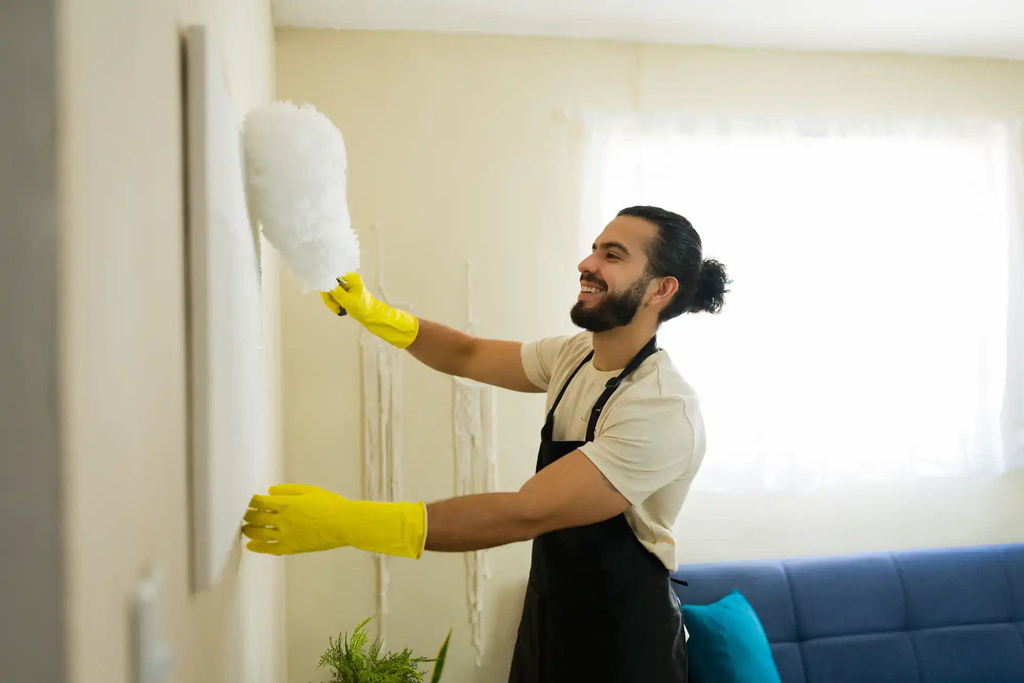 A smiling man wearing yellow rubber gloves and a black apron dusts a picture frame on the wall with a white duster in a bright living room, showcasing the quality of Cleaning Services Long Island, NY.
