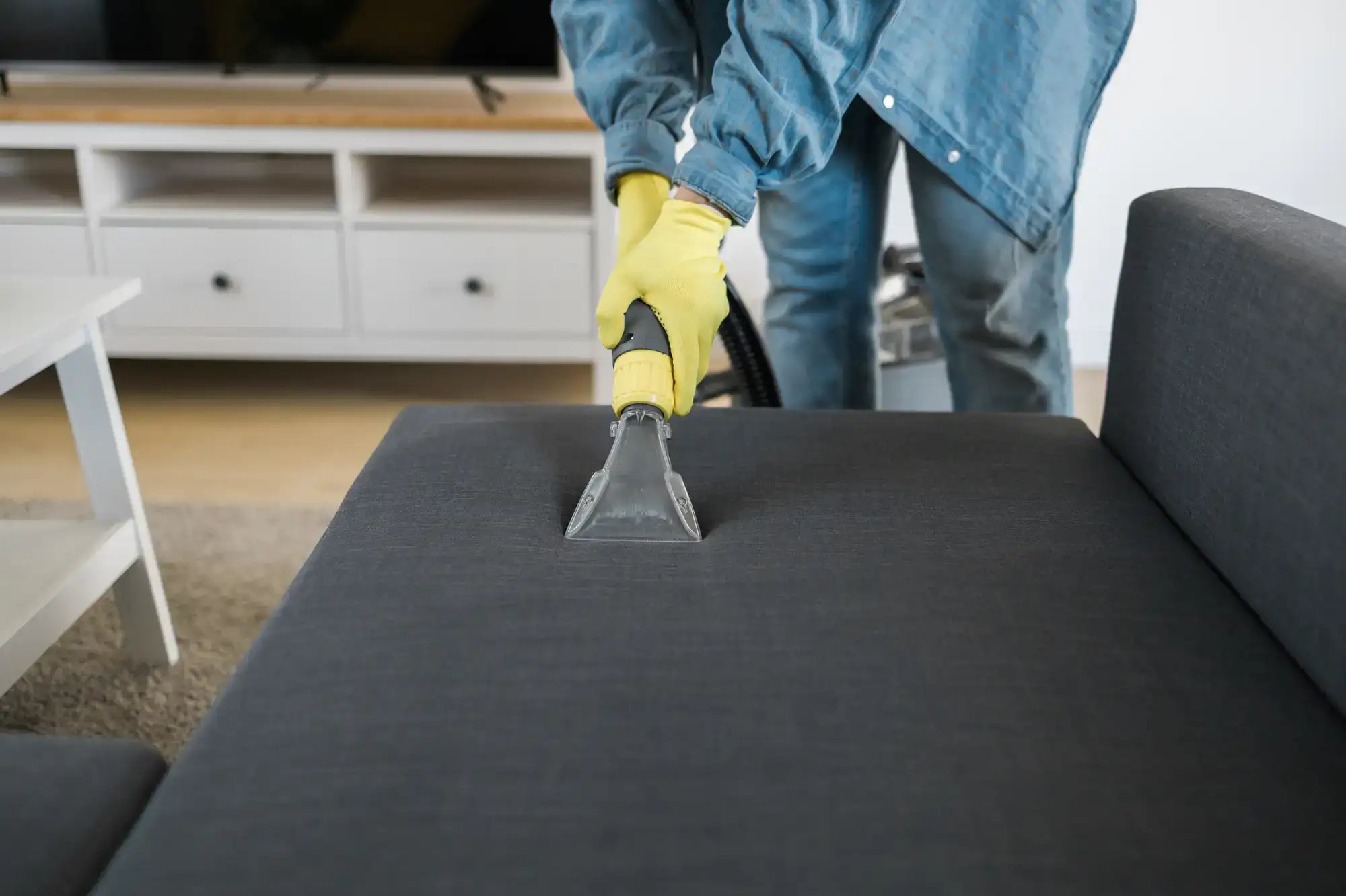 A person wearing yellow gloves uses a vacuum cleaner to clean a dark gray sofa in a stylish NY living room with light furniture and a TV stand—perfect for showcasing expert Cleaning Services Long Island.