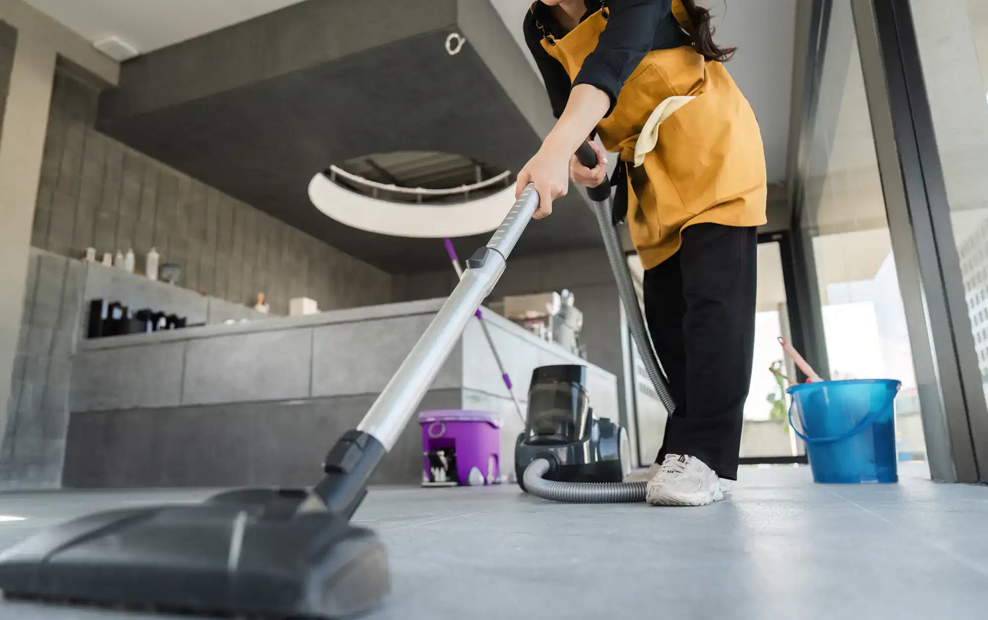 A person wearing a yellow apron vacuums the floor in a modern, bright room, showcasing Cleaning Services Long Island, with cleaning supplies like a mop bucket and purple container visible in the background.