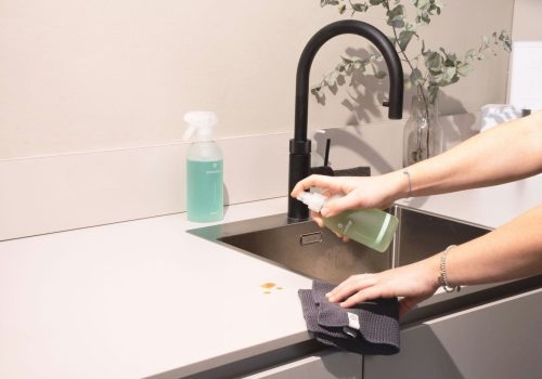 A person sprays cleaner onto a gray cloth near a kitchen sink with a black faucet; showcasing Cleaning Services Long Island, a spray bottle with green liquid and a vase with greenery sit on the countertop.