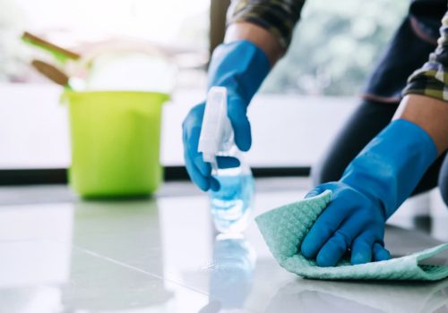 A person wearing blue gloves cleans a shiny floor with a green cloth and spray bottle—typical of NY Cleaning Services Long Island. A green bucket with cleaning supplies is visible in the blurred background.