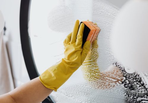 A person wearing a yellow rubber glove is cleaning a soapy glass surface with an orange sponge, showcasing the attention to detail seen in top NY Cleaning Services Long Island.